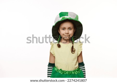 Adorable Irish little child girl wearing clover leaves earrings and green hat, smiles looking at camera, standing isolated over white background. Traditions, culture and national holiday in Ireland