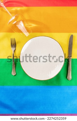 Gay dinner invitation design. A plate with cutlery and a wine glass, overhead flat lay shot on the LGBTQ rainbow flag texture