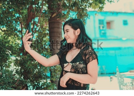 Young fair skinned woman wearing a black and white bikini under a knitted top taking a selfie with her phone.