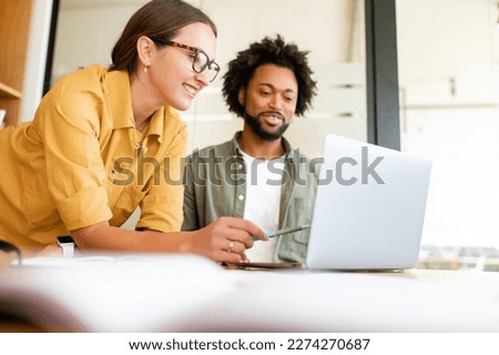 Two colleagues have pleasant conversation sitting side by side in modern open space office in front of laptop, smiling and enjoying friendly working atmosphere, brainstorming together, solving tasks Royalty-Free Stock Photo #2274270687