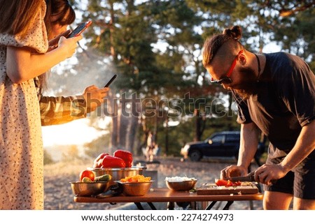 A young man with a beard prepares a salad of vegetables in nature, the girl takes pictures on the phone. Rest friends in the summer in nature, travel, tourism as a hobby, cookout