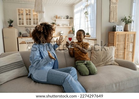 Happy African American family mom and son celebrating Mothers Day together at home, sitting on sofa in living room. Little boy child surprising mum with handmade greeting card. Holiday celebration