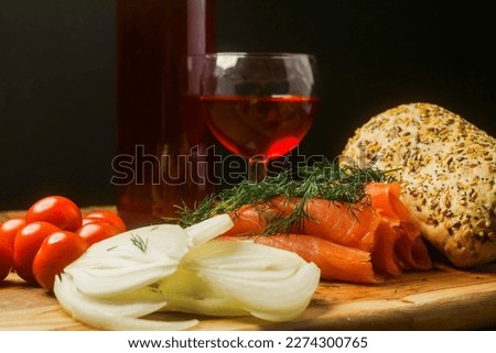 Still life with rolled smoked salmon, red wine, baby tomato, sliced onion and bread roll with seeds on a wooden board, dark background.