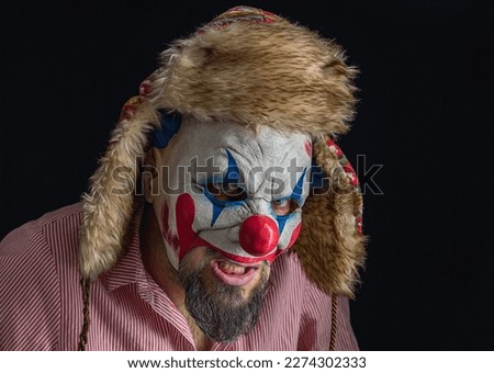 A bearded man wearing a creepy clown mask and a hat with ears looks into the camera. Black background. Scary clown in a red striped shirt grinning in close-up. A maniac clown