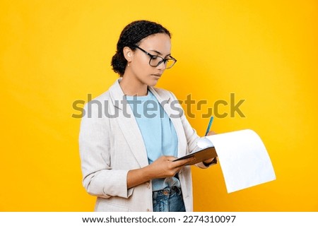 Focused successful brunette mixed race woman, wearing stylish casual clothes, hr manager, real estate agent, holds and sign documents, concentrated looks at them, stand on isolated yellow background