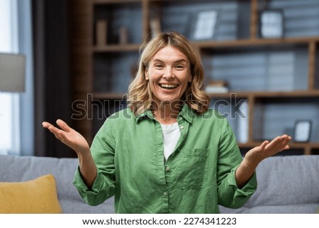 Close-up photo. Portrait of a young happy senior blonde woman sitting on the sofa at home wearing a green shirt. He smiles at the camera, enthusiastically spreads his hands, shows. Royalty-Free Stock Photo #2274341223
