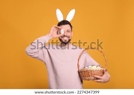 Happy man in bunny ears headband holding wicker basket with painted Easter eggs on orange background