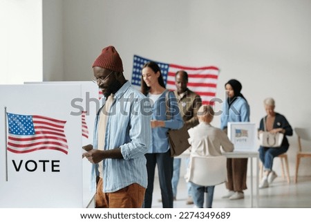 Group of young multicultural voters in casualwear standing in queue along vote booths in polling place and putting their ballots into boxes Royalty-Free Stock Photo #2274349689
