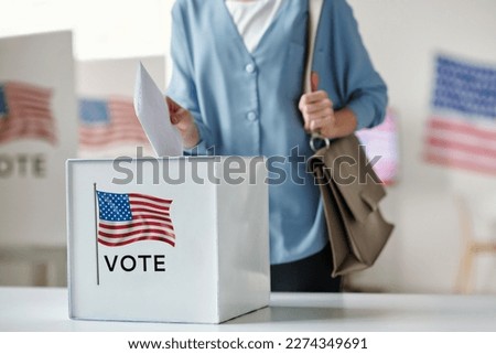 Young female citizen of United States of America putting paper into ballot box while standing by desk in polling place and making her choice Royalty-Free Stock Photo #2274349691