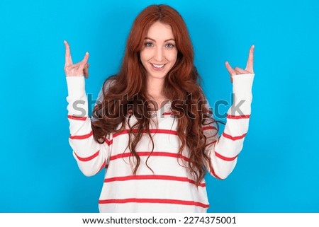 young beautiful red haired woman wearing striped shirt over blue studio background makes rock n roll sign looks self confident and cheerful enjoys cool music at party. Body language concept.