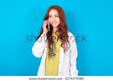 Pleasant looking happy young red-haired doctor woman wearing medical uniform over blue studio background has nice telephone conversation and looks aside, has nice mood and smiles positively while talk