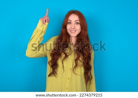 young beautiful red haired woman wearing green shirt over blue studio background pointing finger up and looking inspired by genius thought, showing good idea sign, having clever solution in mind