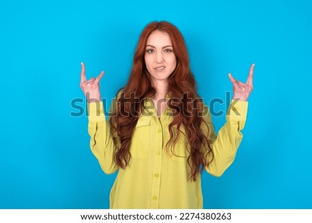 young beautiful red haired woman wearing green shirt over blue studio background making rock hand gesture and showing tongue