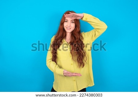 Young red haired woman wearing green shirt over blue background gesturing with hands showing big and large size sign, measure symbol. Smiling looking at the camera.