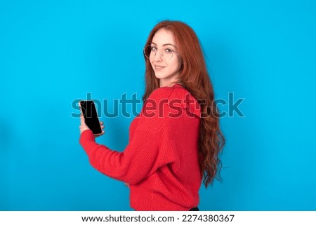 Rear view photo portrait of beautiful red haired woman wearing red knitted sweater over blue background using smartphone smiling