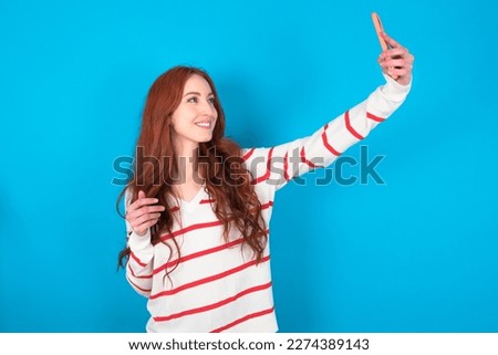 Portrait of a young beautiful red haired woman wearing striped shirt over blue studio background  taking a selfie to send it to friends and followers or post it on his social media.