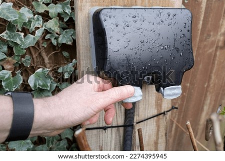 Homeowner about to open the weatherproof cover of a double gang external electrical sockets. Seen in a garden after a heavy downpour. Royalty-Free Stock Photo #2274395945
