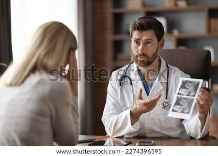 Male Gynecologist Doctor Consulting Pregnant Female Patient In Clinic, Professional Reproductive Endocrinologist Showing Baby Sonography Image To Stressed Lady, Discussing Screening Result
