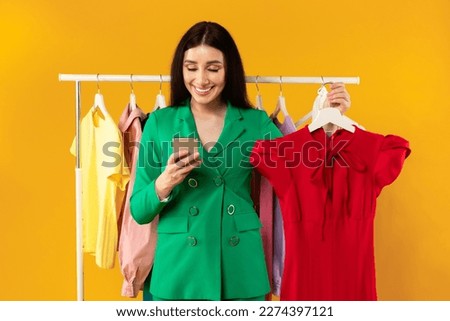 Mobile shopping concept. Happy lady using cellphone and holding hanger with new red dress, ordering summer fashion trends on yellow background. Fashion and style app for smartphone Royalty-Free Stock Photo #2274397121