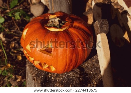 An orange pumpkin with an evil face carved on it for Halloween and an ax lie on a wooden stump.