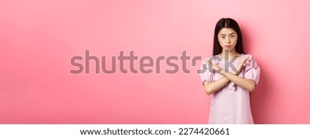 Serious chinese girl show cross sign, make stop gesture and look confident, block person, say no and reject offer, standing on pink background.