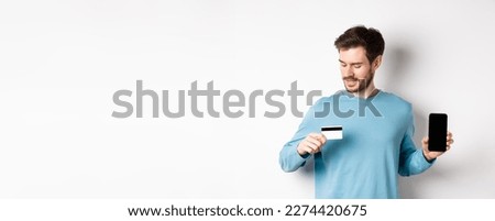 Handsome young man with beard, showing empty smartphone screen and looking at plastic credit card, standing over white background.