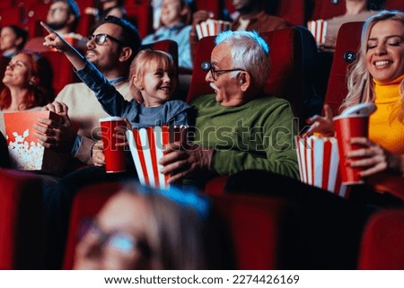 A senior Caucasian man is with his little granddaughter watching a movie in the cinema. They are bonding over the experience with popcorn as she's pointing to the big screen to him. Royalty-Free Stock Photo #2274426169