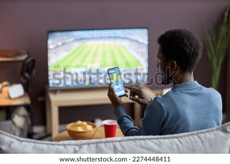 Back view of man holding smartphone with sports bets app on screen while watching football match at home, copy space Royalty-Free Stock Photo #2274448411