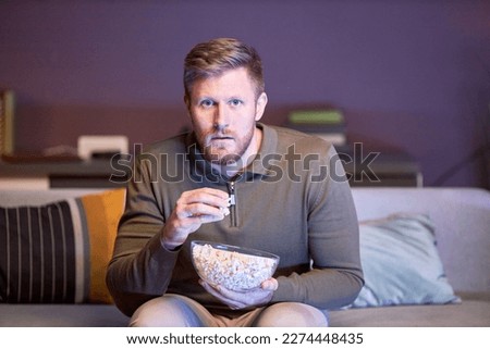 Portrait of one man watching sports match on TV at home in blue light and eating popcorn, copy space