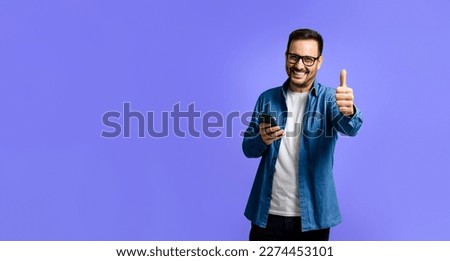 Portrait of cheerful young adult man in denim shirt using smart phone and showing thumbs up sign while standing isolated over blue background