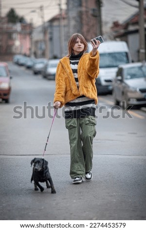 Young pretty woman in a yellow fur coat, social media influencer, walk the streets of the city with her black dog, using smartphone.