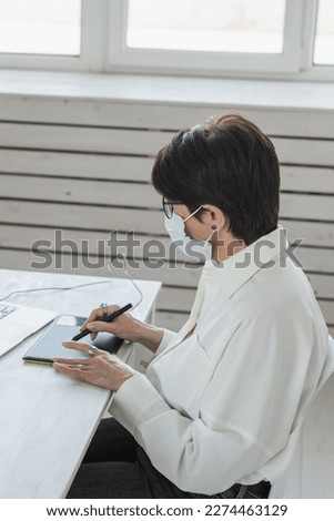 Middle aged woman in medical face mask graphic designer or photographer using digital graphic tablet while working at modern office, professional female retoucher sitting at modern workspace, generic