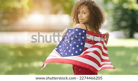 Young African american woman at nature background with an American USA flag on her shoulders. USA resident, US citizen. Immigration concept                               