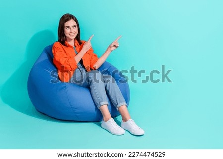 Full length photo of dreamy shiny lady wear orange shirt sitting bean bag pointing empty space isolated teal color background
