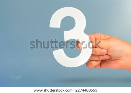 Number three in hand. Hand holding white number 3 on blue background with copy space. Concept with number three. 3 percent, birthday 3 years, third grade, triple Royalty-Free Stock Photo #2274480553