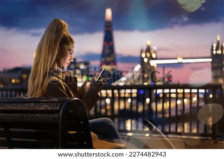A urban woman sitting on a bench and looking at her smartphone during night time in front of the illuminated city skyline of London, England