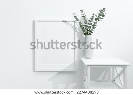 Blank white frame mockup in white room interior with fresh eucalyptus plant decor, copy space