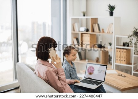Back view of woman ordering pizza via smartphone while sitting with little girl on sofa. Pleasant mother and cute daughter spending quality time together while buying takeaway lunch with laptop.