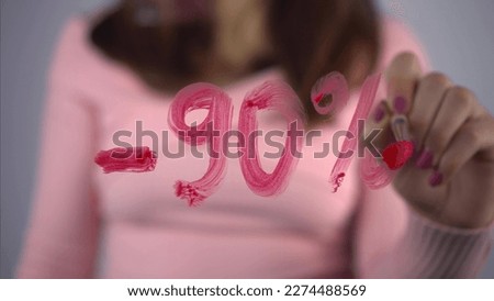 A young woman draws on the glass discounts minus 90 percent. A girl in a pink sweater draws with a brush and red paint. Close-up.