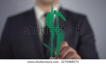 A young businessman draws a dollar symbol on a transparent glass. A man draws with green paint and a brush close-up.