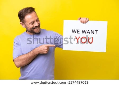 Middle age caucasian man isolated on yellow background holding We Want You board and  pointing it
