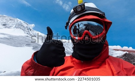 Climber with helmet, headlamp and black jacket taking a selfie on a glacier on a cloudy day, landscape reflected in red framed glasses
