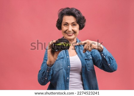 Mature senior woman eating burger with satisfaction. Grandmother enjoys tasty hamburger takeaway, delicious bite of burger, order fastfood delivery while hungry, isolated over pink background.