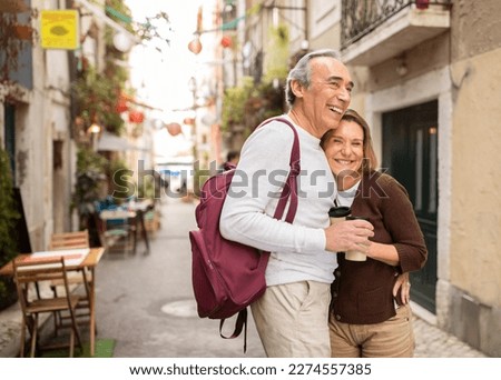 Joyful Senior Tourists Couple Hugging Posing With Backpack Holding Paper Coffee Cups On Cozy Lisbon Street With Outdoor Cafes. Retired Elderly Spouses Enjoying Vacation And Retirement In Europe Royalty-Free Stock Photo #2274557385