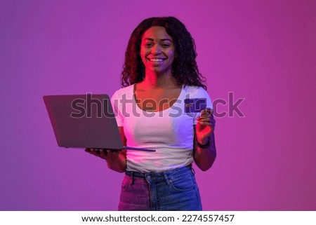 Online Payments. Smiling african american woman with credit card and laptop in hands standing in neon lighting over purple background, happy young black female making internet shopping or paying bill