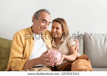 Happy european senior couple holding pink piggybank in hands and embracing, sitting on sofa in living room interior, free space. Retirement savings concept Royalty-Free Stock Photo #2274557611