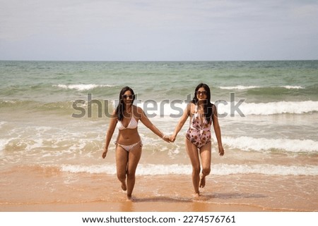 Two beautiful young women in bikinis strolling along the shore. The women are enjoying their trip to the beach paradise while making different gestures and expressions. Holidays and travels.
