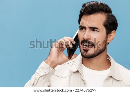 Close-up portrait of a brunette man talking on the phone, disgruntled and angry, on a blue background in a white T-shirt and jeans, copy space