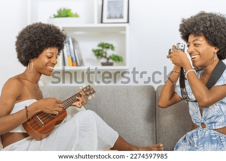 Identical black twin sisters with afro hairstyles, one playing a ukulele and the other taking pictures of her sister with an antique camera, sitting on the couch in the living room of their apartment.