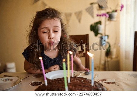 Authentic portrait of a Caucasian child, sweet baby girl making cherished wish while blowing out colorful lit candles on her birthday cake. Happy kid girl celebrates her 5th birthday at home